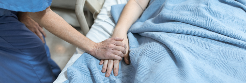 Potilas makaa peiton alla sairaalasängyssä ja hoitaja pitelee hänen kättään.