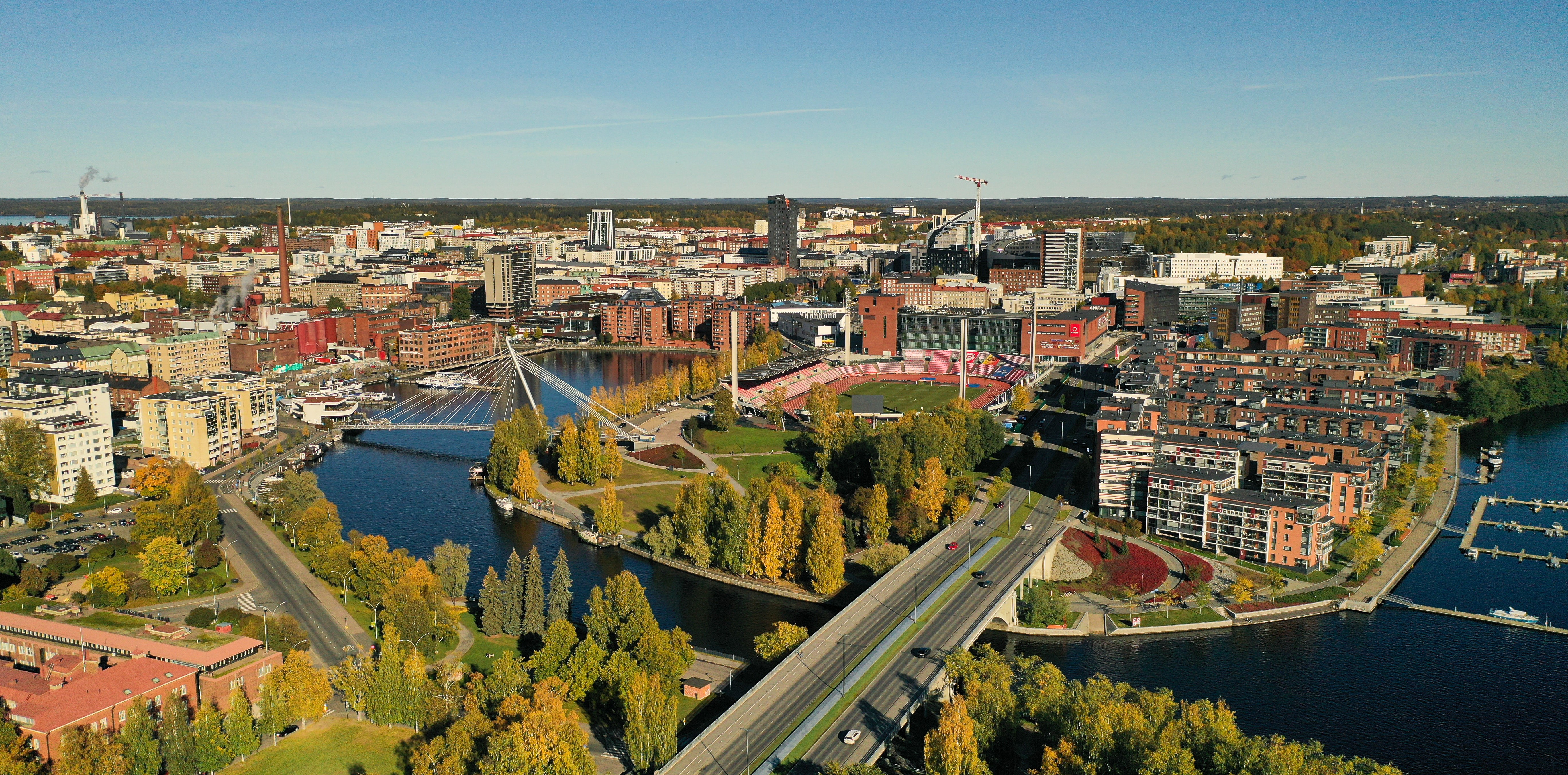 Tampereen keskustan etualalla näkyy Ratinan silta, joka ylittää veden. Sillan takana näkyy kävelysilta ja Ratinan suvanto ja Ratinan stadion. Lisäksi kuvassa näkyy paljon kerrostaloja ja muuta kaupunki-infrastruktuuria.