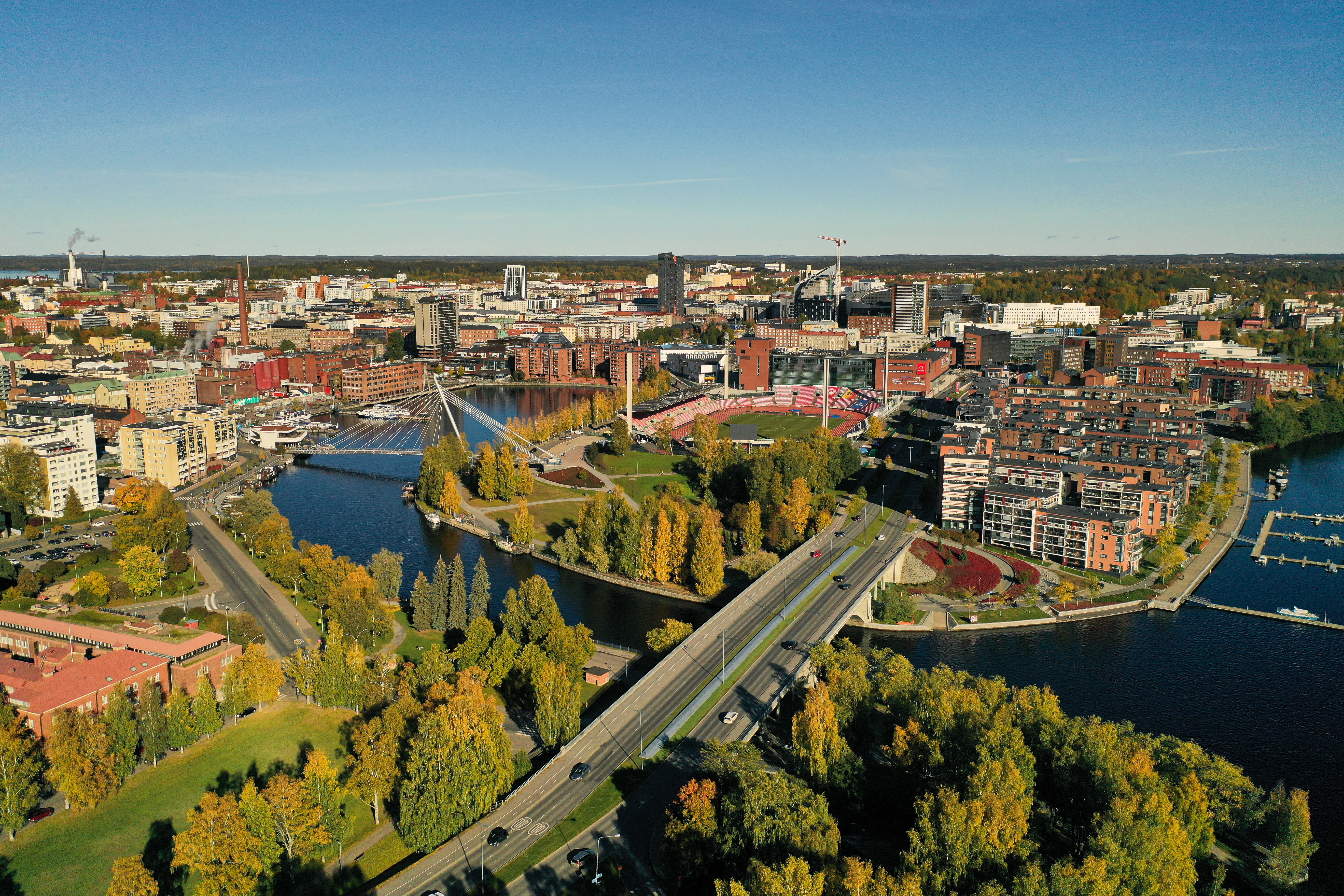Tampereen keskustan etualalla näkyy Ratinan silta, joka ylittää veden. Sillan takana näkyy kävelysilta ja Ratinan suvanto ja Ratinan stadion. Lisäksi kuvassa näkyy paljon kerrostaloja ja muuta kaupunki-infrastruktuuria.