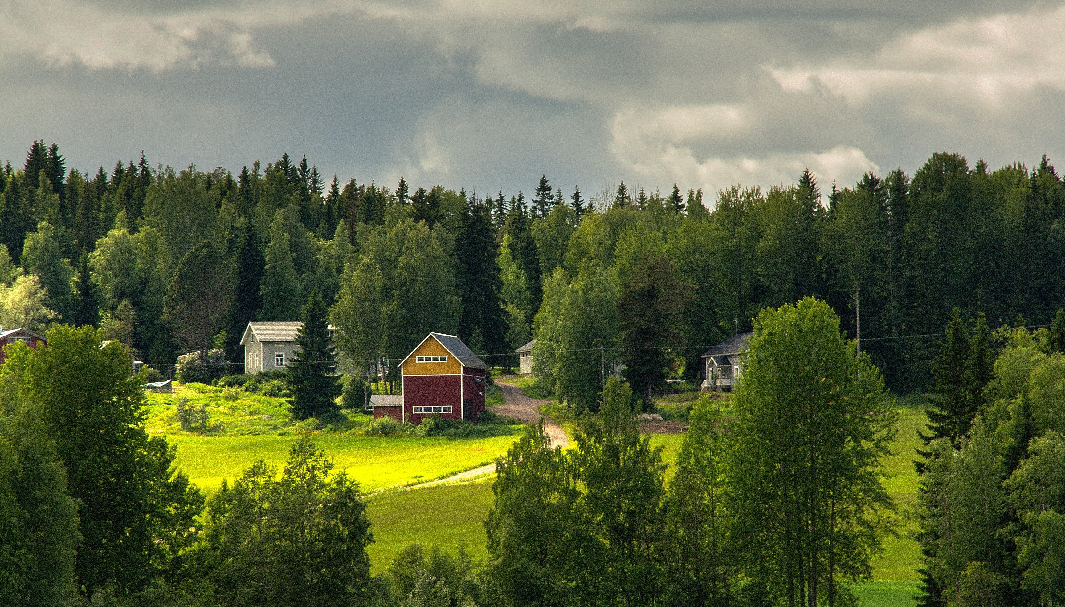 Maalaismaisemassa näkyy kesäinen vihreä pelto, jonka ympärillä kasvaa harvakseltaan puita. Pellon keskellä kulkee kapea hiekkatie, joka johtaa pellon takana olevien talojen luo. Talot ovat puisia ja harjakattoisia. Näkyvimmin etualalla on vanha rakennus, jonka alaosa on tummanpunainen ja yläosa tummankeltainen.