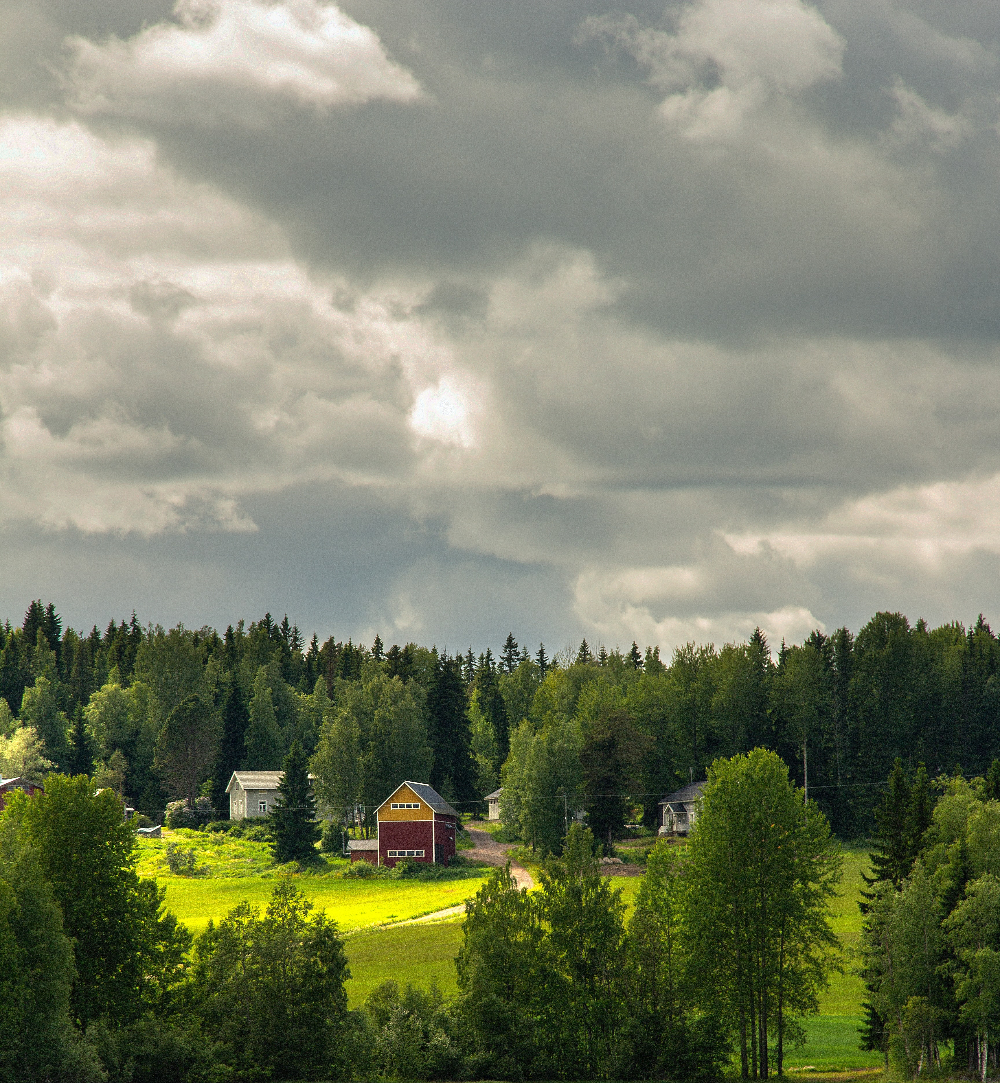 En sommargrön åker i en lantlig miljö och kring den växer några träd. Mitt i åkern löper en smal sandväg som leder till några hus bortom åkern. De är trähus med sadeltak. I förgrunden syns en gammal byggnad med mörkröd nedre del och mörkgul övre del. 