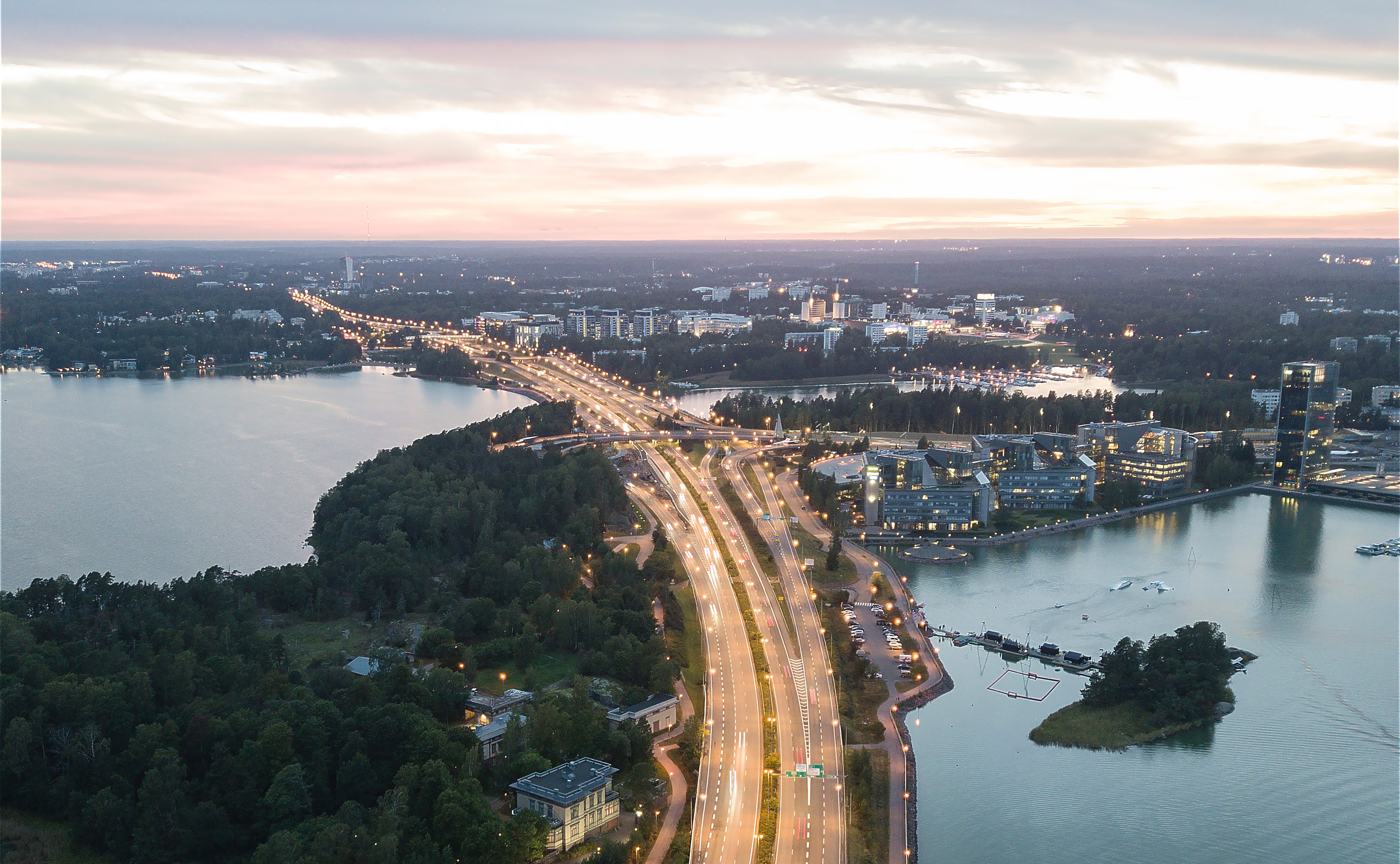 Veden ylittävä moottoritie on valaistu katuvaloilla. Tien ympärillä näkyy veden lisäksi kaupunkia kerrostaloineen. On ilta, mutta ei ole vielä täysin pimeää. Taivaanrannasta kajastaa vielä valoa.