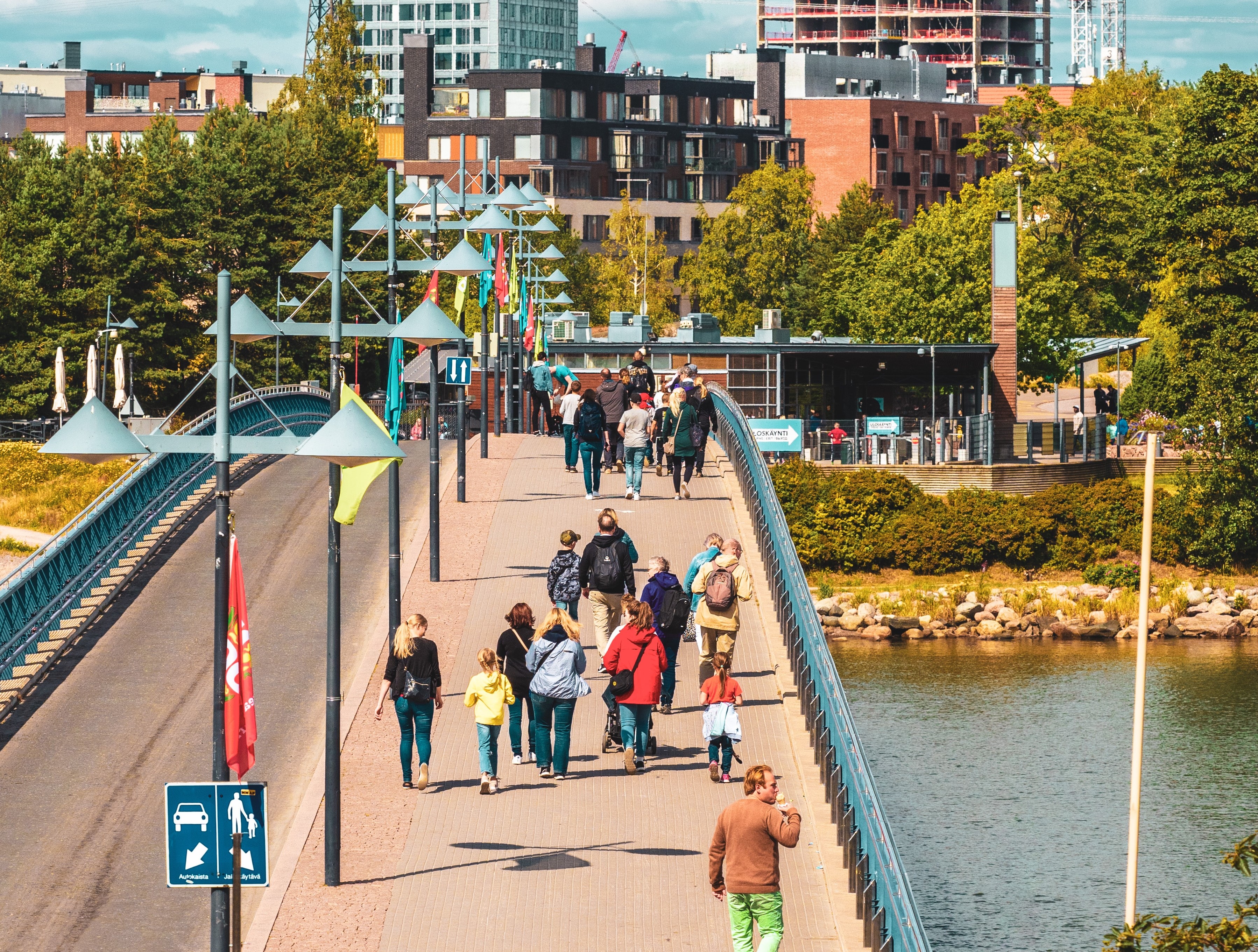 Ihmiset kävelevät sillalla, joka ylittää veden. Takana näkyy korkea pilvenpiirtäjä ja sen vieressä rakenteilla oleva korkea talo ja kaksi nosturia.