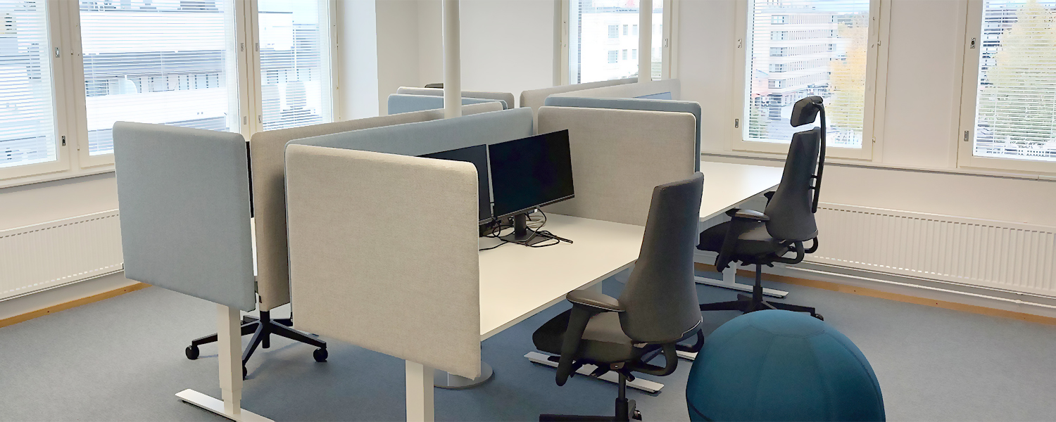 Fyra skrivbord i ett öppet kontorslandskap. Skrivborden är placerade 2 och 2 mot varandra. Mellan skrivborden finns gråa och ljusblåa skärmar. Varje skrivbord har två skärmar och framför varje bord finns en kontorsstol. Bakom skrivborden finns fönster, utanför syns höghus.