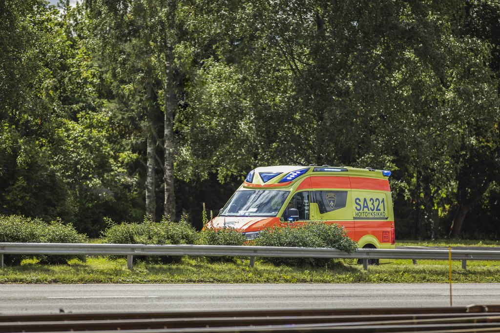 En ambulans kör längs en landsväg. Det är sommar och bakom ambulansen syns gröna lövträd. 