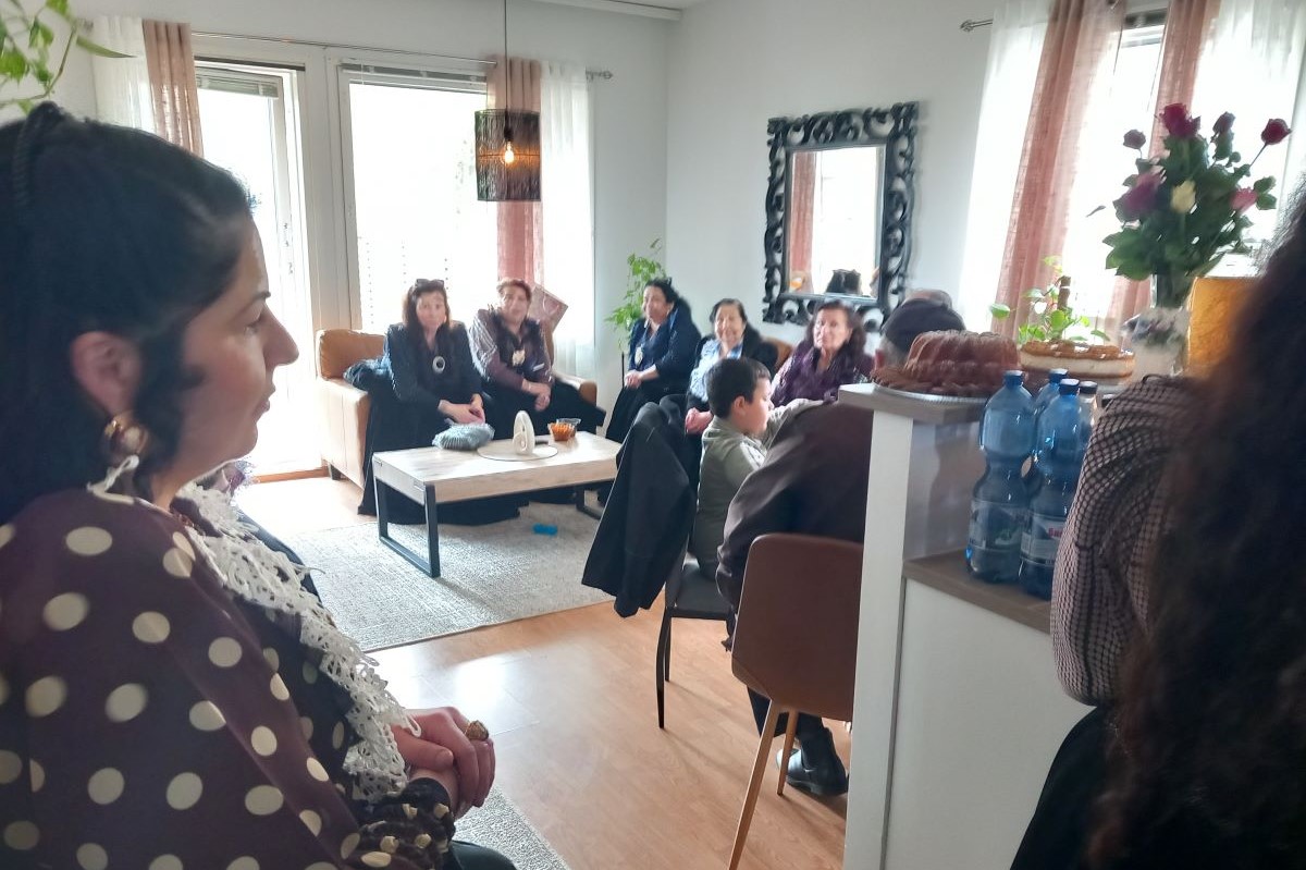 Romanipukuisia henkilöitä asunnossa. Kaksi henkilöä näkyy etualalla ja useampi henkilö istuu taka-alalla huoneessa.