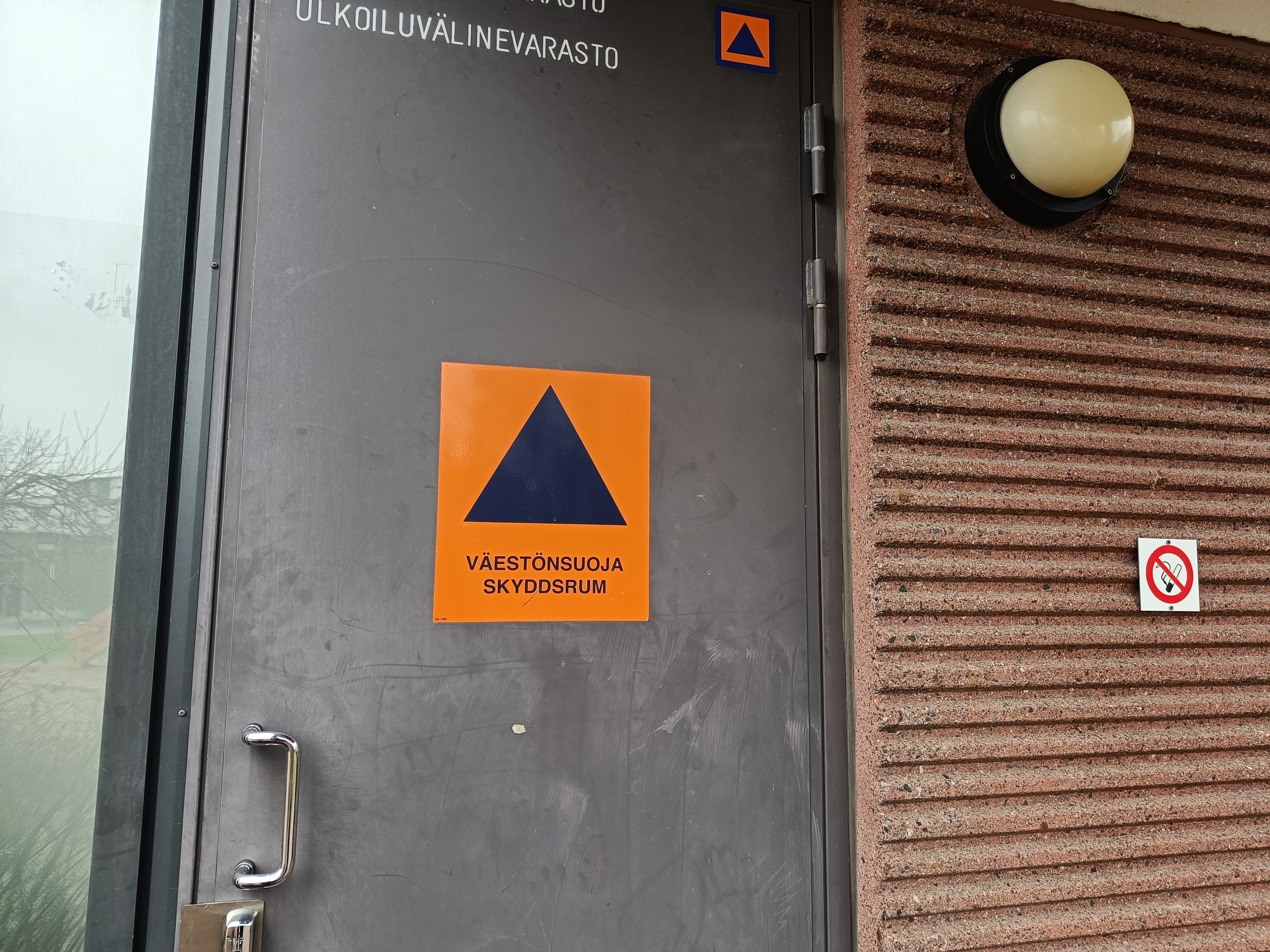ovi kerrostalon ulkoiluvälinevarastoon, joka toimii myös väestönsuojana. Ovessa on väestönsuojan kansainvälinen logo, jossa on tummansininen kolmio oranssilla pohjalla ja alla teksti väestönsuoja skyddsrum.