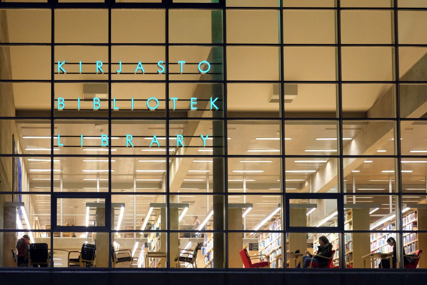 Library photographed from the outside. You can see people sitting and reading behind the glasses, in the well lit library.