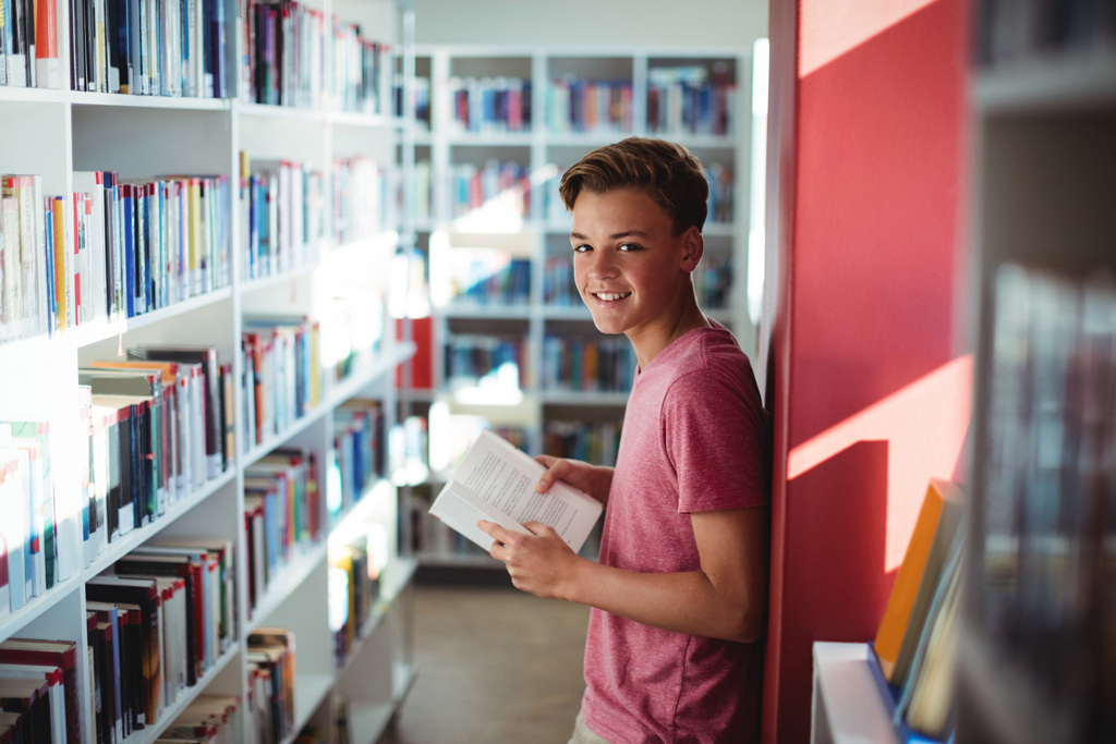 Hymyilevä poika, joka seisoo kirjastossa kirja kädessä.