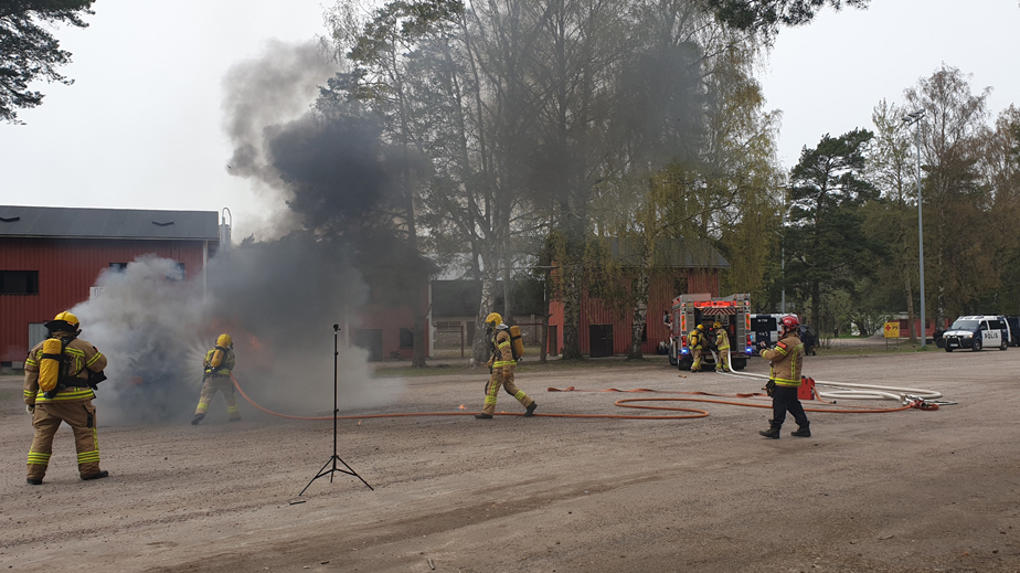 Pelastustoimen harjoitus, jossa pelastajat sammuttavat savuavaa tulipaloa. Taustalla paloauto ja poliisiauto.